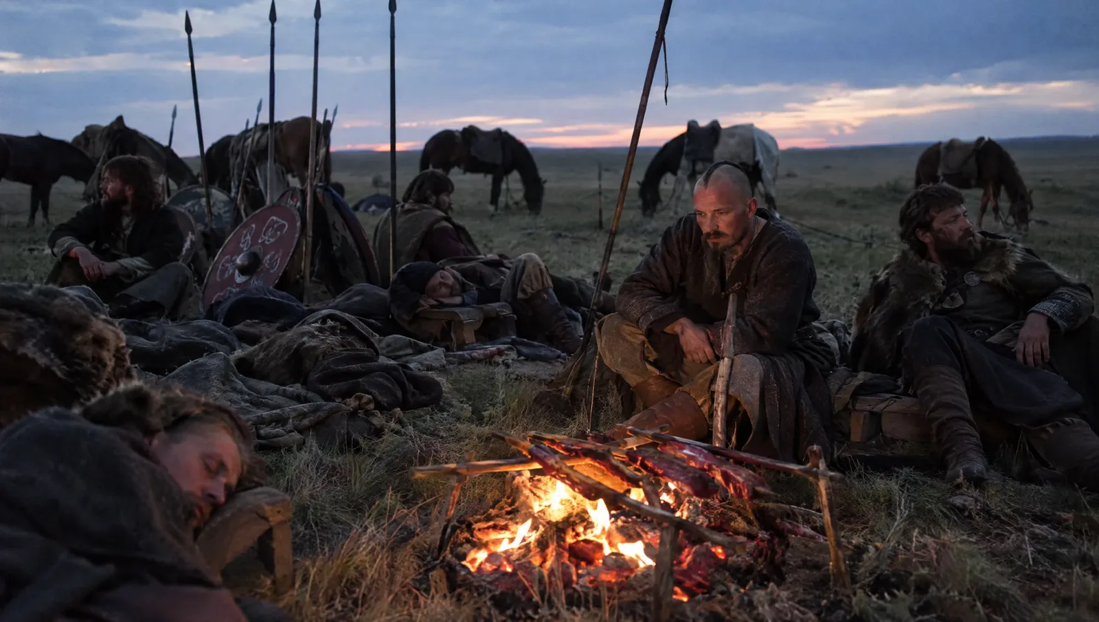 Rus' warriors resting on horse-blankets with saddles as pillows and roasting strips of meat on coals on the steppe at dusk